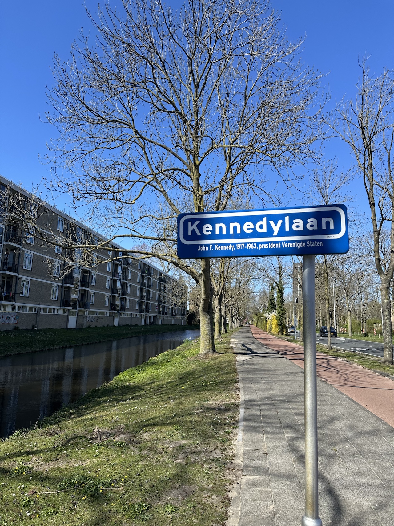 A Dutch street sign reading 'Kennedylaan' beside a tree-lined canal and a red-paved cycle path separated from the road