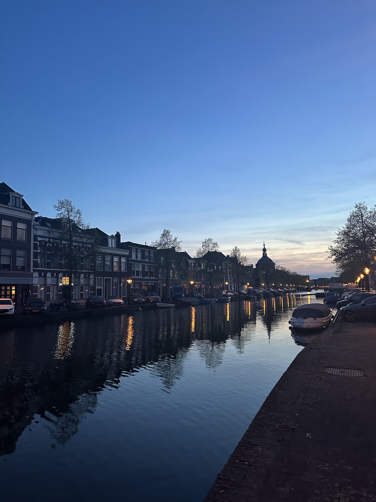 A Leiden canal at dusk, lined with brick townhouses and moored boats, with a domed church silhouetted in the distance