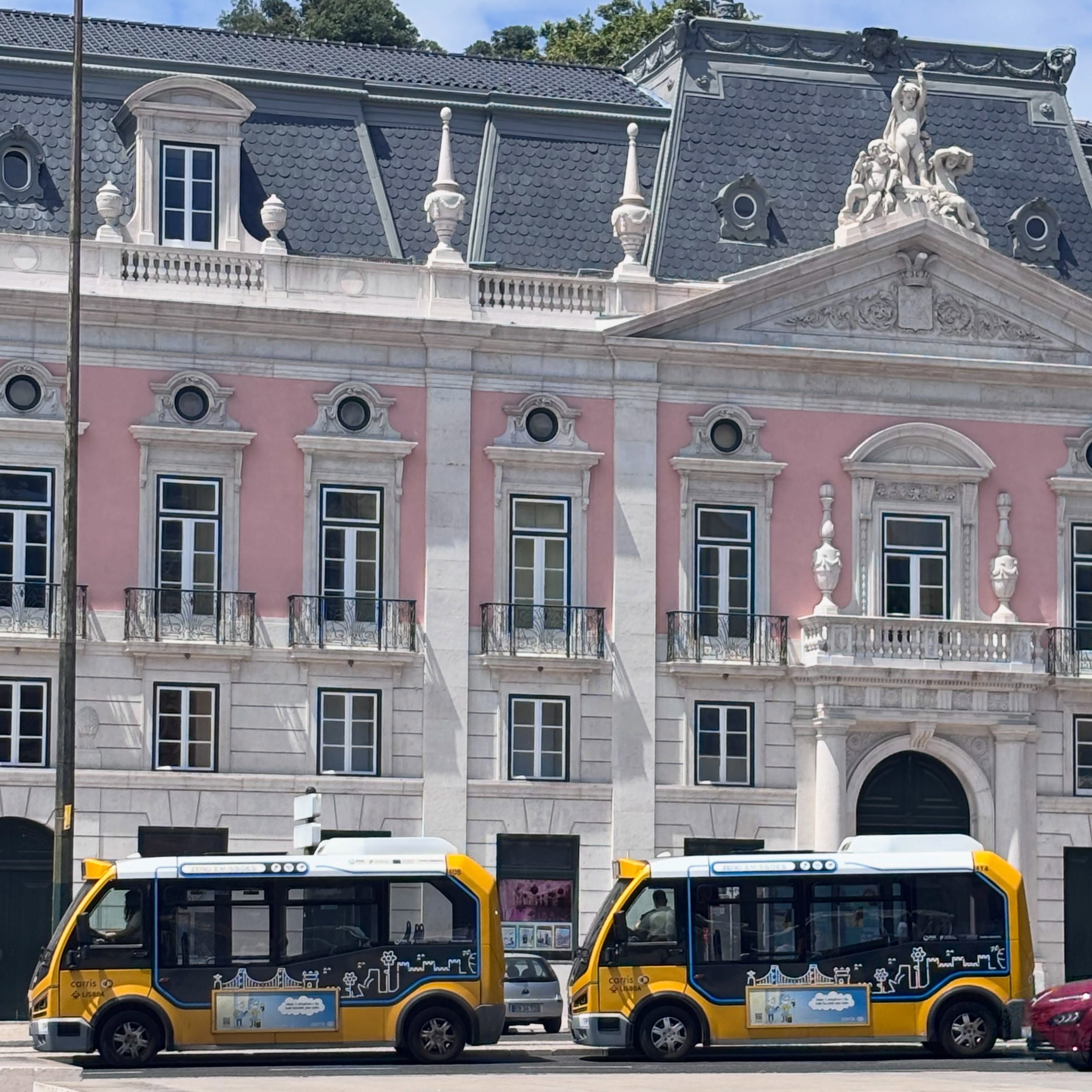 A typical Lisbon building, pink, with two KARSAN e-Jest mini public transit buses stopped in front