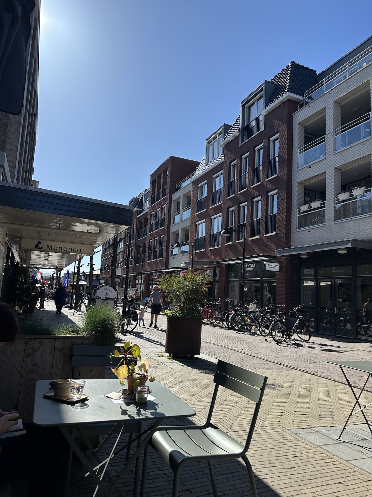 A sunlit pedestrian street in Voorschoten with café tables in the foreground, brick apartment buildings, and rows of parked bicycles